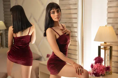 Young woman in a burgundy velvet mini dress posing in a warm, elegant bedroom setting.