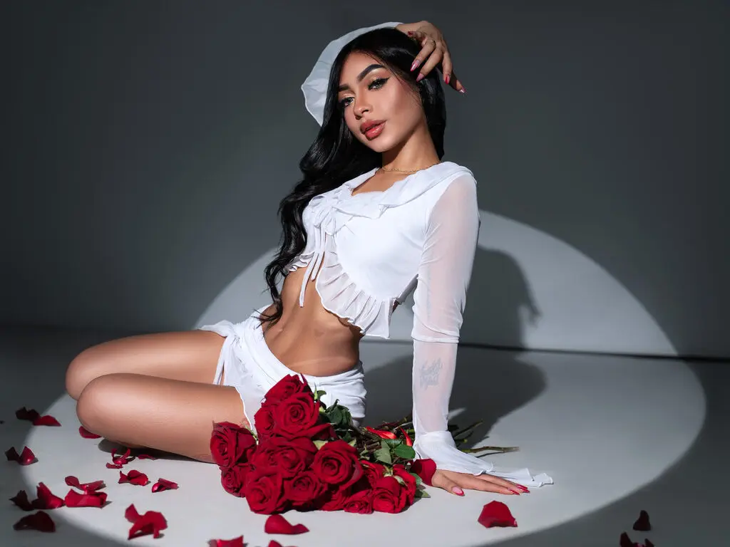 Glamorous woman in a white crop top and shorts sitting by red roses in a moody studio setting.