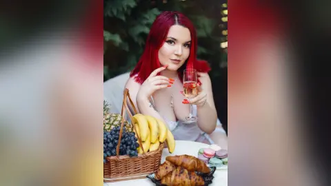 Red-haired woman in white lace enjoying champagne with a fruit basket and macarons in a bright outdoor setting.