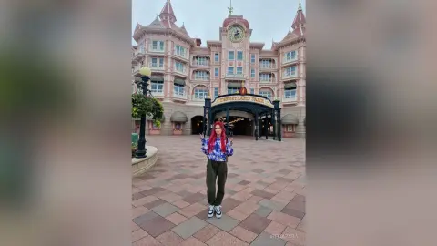 Red-haired woman in a blue patterned jacket and dark trousers posing happily in front of Disneyland Park.