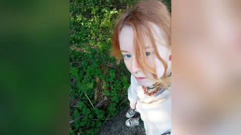 Redheaded woman in a white top and beaded necklace relaxing in a lush green forest setting.