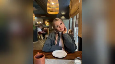 KrystalLaaz sits at a restaurant table with her blonde hair in braids, wearing a grey zip-up sweater.
