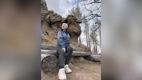Smiling woman in a blue jacket and beanie sitting on a log near rocky cliffs in a serene forest setting.