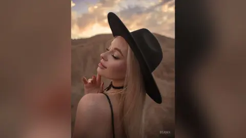 BeaNix poses outdoors in a black hat and black choker, looking back over her shoulder with long blonde hair.