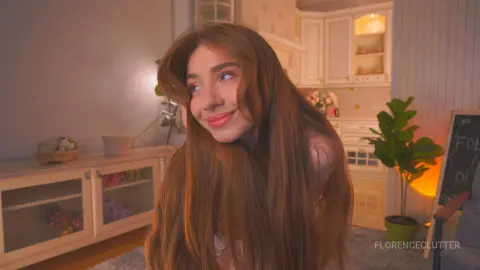Smiling young woman with long brown hair in a cozy, warm-lit aesthetic room.