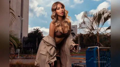 Stylish blonde woman in a beige trench coat and lace lingerie posing outdoors in a sunny urban setting.