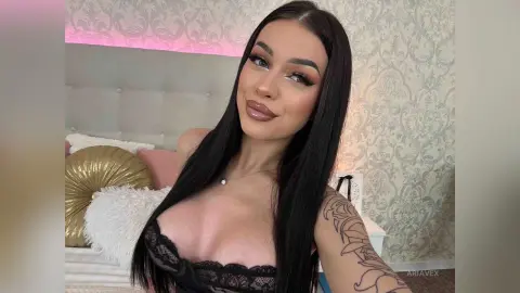 AriaVex poses in a bedroom wearing black lace lingerie, showing her long black hair and shoulder tattoo.