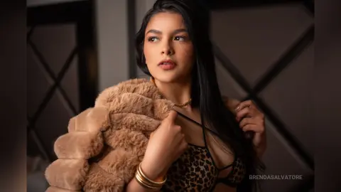 Beautiful woman in a leopard print bikini and tan faux fur coat posing in a moody indoor setting.