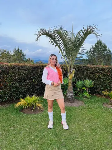 Ninablaze27 stands in a garden wearing a pink vest, white shirt, and tan skirt with tall white socks.