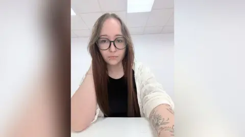 LizbethLocantore wearing glasses and a black top with a white open shirt, showing her long brown hair and arm tattoos.
