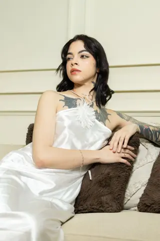 Tattooed woman in a white satin slip dress relaxing on a couch in a bright, minimalist room.