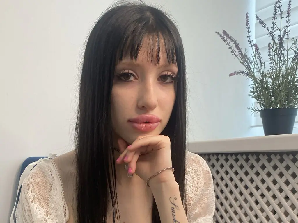Young woman with dark bangs in a white lace top posing thoughtfully in a bright indoor setting.