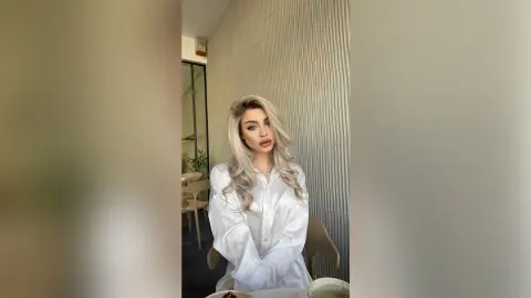 Blonde woman in a white silk blouse posing in a modern minimalist office setting with a sophisticated mood.