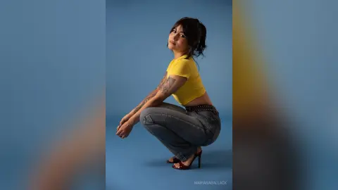 Young woman in yellow crop top and grey jeans posing confidently against a blue studio background.
