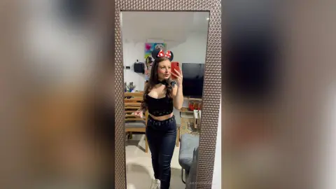 Young woman in black crop top and jeans wearing Minnie Mouse ears taking a selfie in a bedroom mirror.