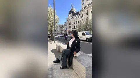 NashiroSakurai sits on a stone ledge wearing a black coat and grey trousers, looking off to the side against a city street background.