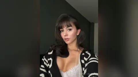 Young woman with brunette bangs wearing a white lace top and patterned cardigan in a moody indoor setting.