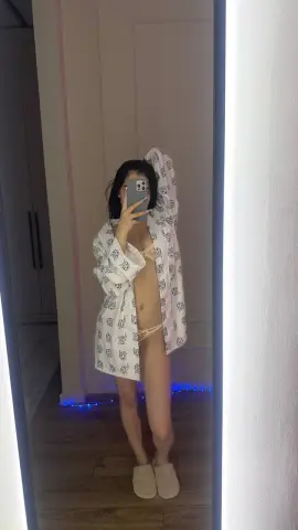 Young woman in a white patterned robe and beige bikini posing for a mirror selfie in a modern bedroom.