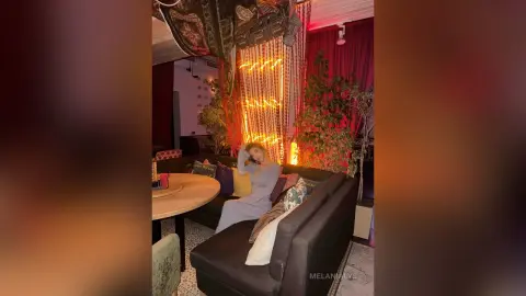 Woman in a grey dress relaxing on a sofa in a cozy, dimly lit room with warm fairy lights and bohemian decor.