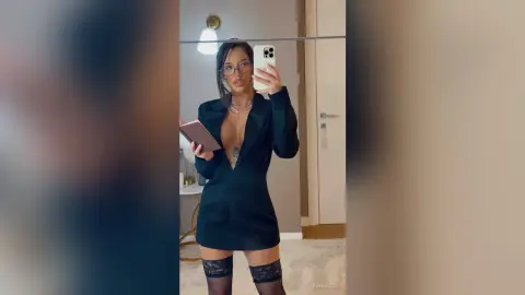 Young woman in a black blazer dress and glasses taking a mirror selfie in a modern bedroom.