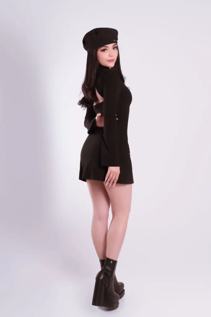 EmmaWeest in a black long-sleeve mini dress and beret poses on her hands and knees against a white background.