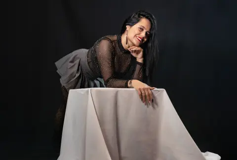 Smiling woman in a sparkly black sheer top and grey skirt posing against a dark studio background.