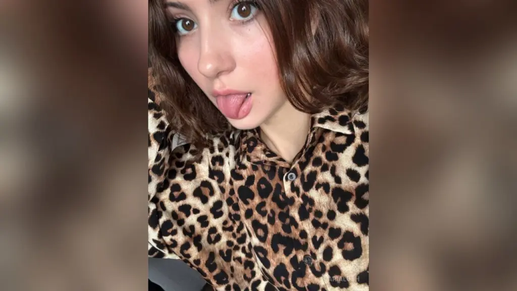 Young woman with lip piercings and a black lace choker making a peace sign in a casual indoor setting.