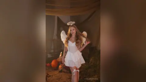 StellaFletcher wears a white lace angel outfit with wings and a halo while standing in a dim, warm setting.