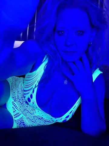 Blonde woman in a white lace bodysuit posing under moody blue neon lighting.