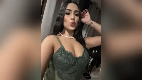 Young woman in a green lace camisole making a playful kissy face in a dimly lit indoor setting.