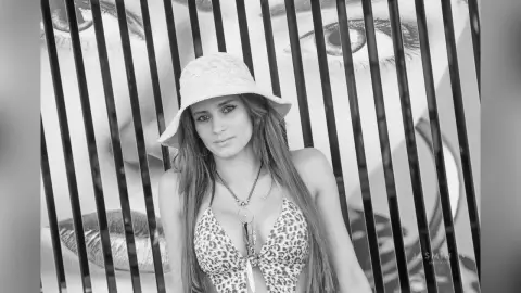 AndreaKalo wears a white sun hat and a leopard print bikini top, posing in front of a large black and white face graphic behind vertical bars.