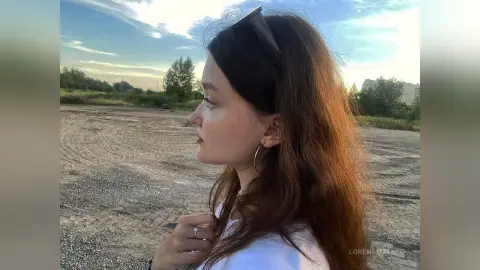 Side profile of a young woman with long brown hair looking away in a dusty outdoor field at sunset.