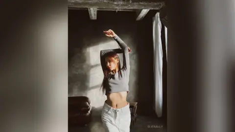 Young woman in a grey long-sleeve crop top and white jeans posing in a moody, sunlit industrial loft.
