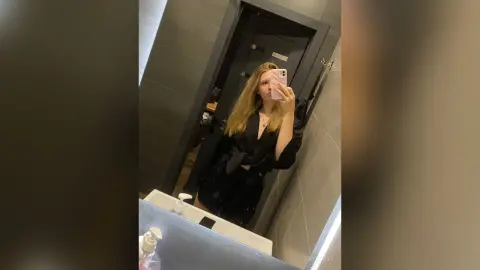 Young woman in a black satin outfit taking a mirror selfie in a modern bathroom setting.