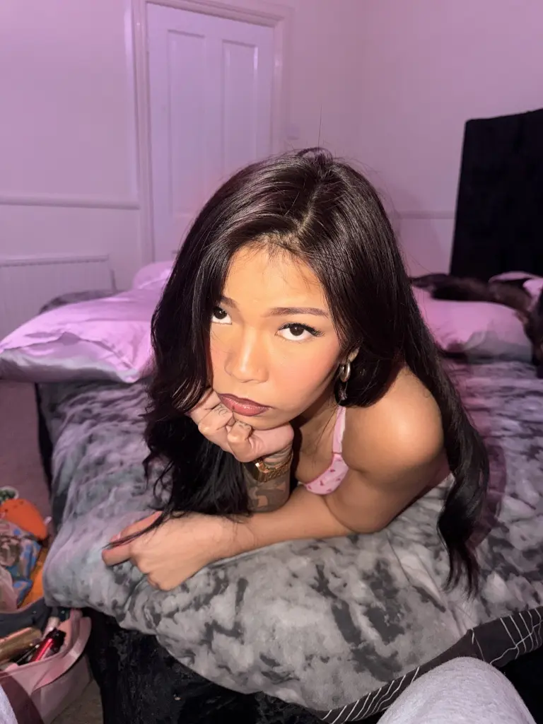 BabyDollJaz poses with her hands covering her mouth, showing her long dark hair and bare shoulders while lying on pink bedding.
