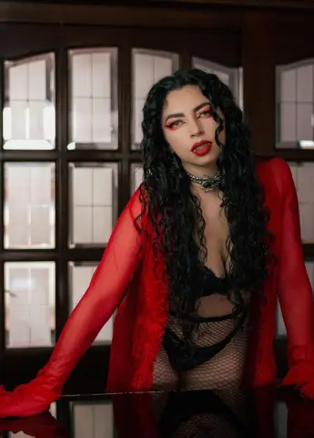 Woman with long curly hair in a red sheer cardigan and black lace lingerie posing in a moody indoor setting.