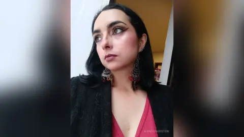 Woman with dark hair and heavy eyeliner wearing large silver ethnic earrings and a black blazer in a soft indoor light.