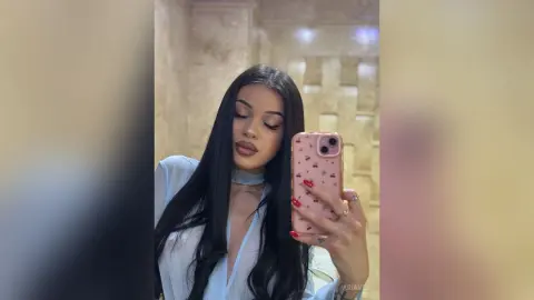 AriaVex looks into the camera while holding a pink patterned phone, wearing a light blue top with a cutout neck.