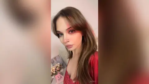Young woman with long brown hair and red eye makeup wearing a red lace top in a soft, romantic indoor setting.