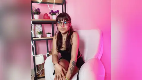 Young woman with glasses in a black tank top sitting in a white chair against a pink aesthetic background.