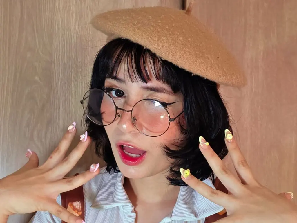 Trendy woman with short dark hair, round glasses, and a tan beret posing with colorful manicured nails.