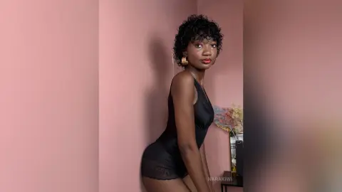 Young woman in a black bodysuit posing against a pink wall in a moody, minimalist indoor setting.