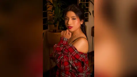 NairaWestbrook poses in a red plaid shirt falling off her shoulders, showing her bare shoulders and neck, with long dark hair and red lipstick.
