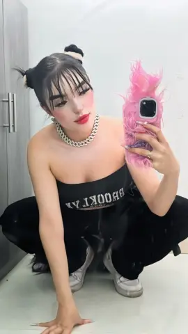 Young woman with space buns in a black tank top taking a mirror selfie in a modern room.