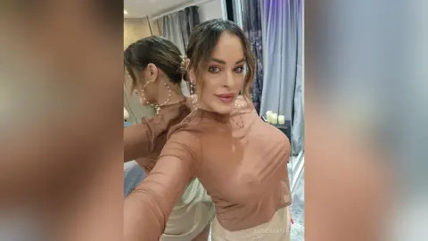 Glamorous woman in a sheer brown long-sleeve top and gold jewelry posing for a selfie in a modern room.