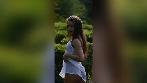 Dreamy portrait of a girl in a white dress and flower crown standing in a lush green forest.