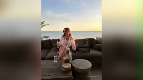 Stylish woman in a white linen outfit relaxing on a lounge sofa by the ocean at sunset.
