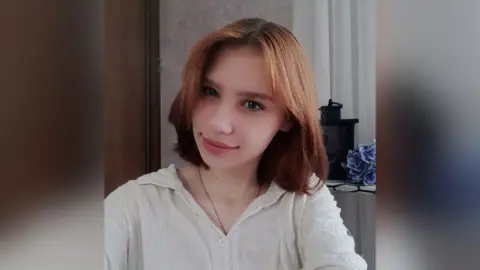 Smiling young woman with reddish-brown hair wearing a white linen shirt in a bright indoor setting.