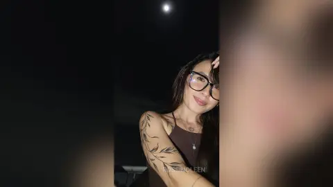 RossiNoleen poses wearing glasses and a dark brown tank top, showing a leaf tattoo on her shoulder against a dark night sky.