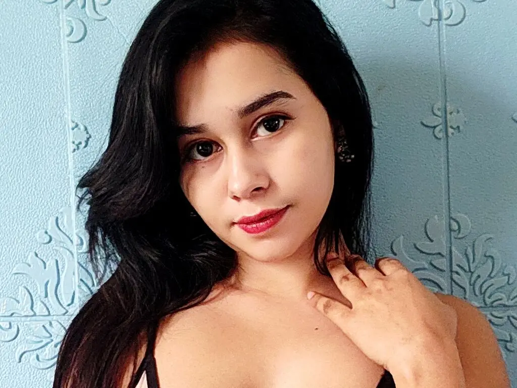 Young woman with long dark hair posing in a black top against a light blue textured wall background.